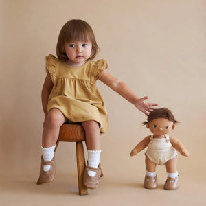 A DINKUM DOLL from OLLI ELLA USA features soft brown hair and comes dressed in a white knit romper paired with brown shoes. This charming doll has an embroidered face with brown eyes, a small nose, and a gentle smile. Its posable body makes it perfect for imaginative playtime, and it is posed standing upright.