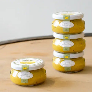 A small, transparent jar of "BELLA CUCINA - GOLDEN PEPPER PESTO" with a white lid. The jar contains a thick, yellow-colored sauce and features a light green label on the front displaying the product name and weight (6 oz. / 169 g). This vegan, small-batch delicacy from BELLA CUCINA is placed on a clean white surface.