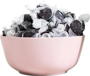 A light pink bowl is filled with numerous individually wrapped pieces of JACOBSEN SALT - SALTY BLACK LICORICE. The wrappers, twisted at both ends, partially expose the dark candy inside. The white and dark contrast of the wrappers complements the pastel color of the bowl.