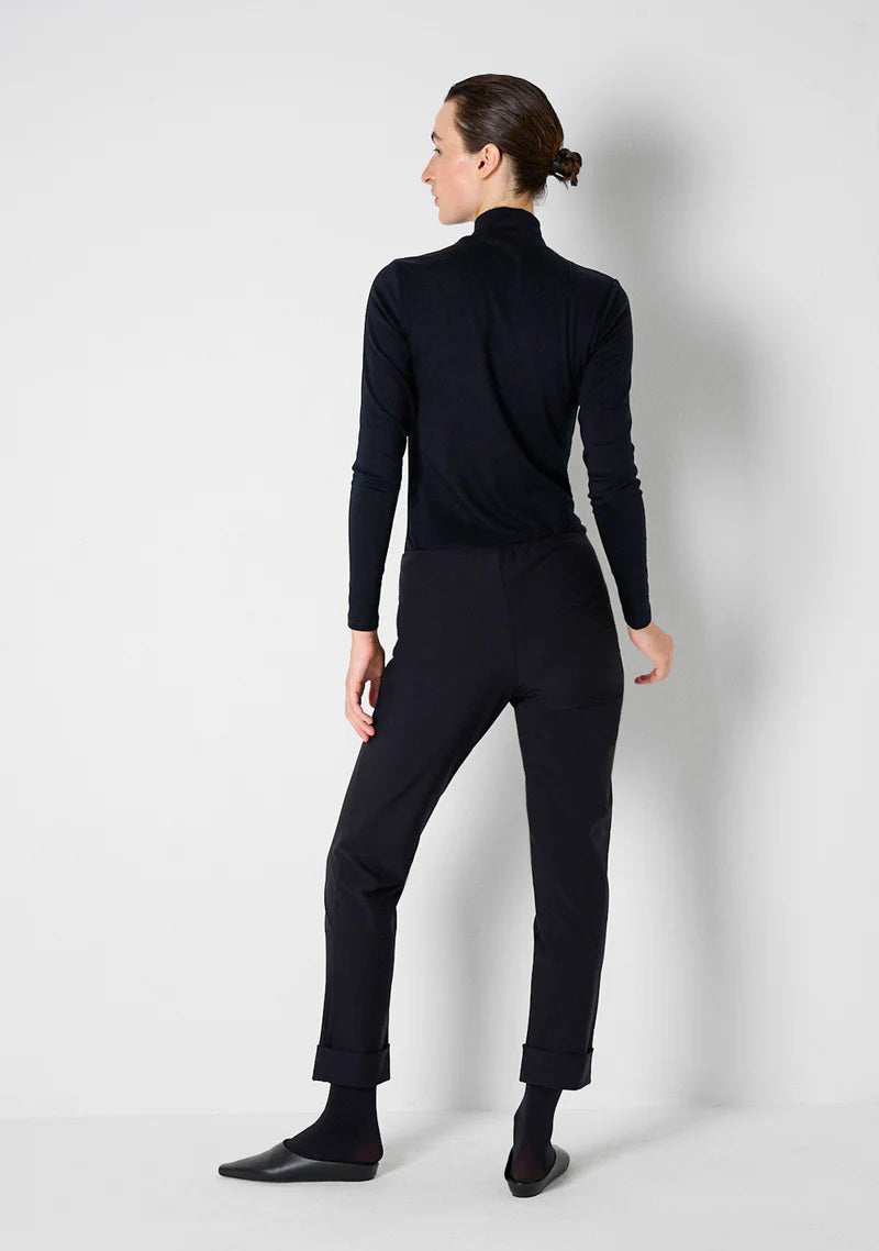 A person in KATHARINA HOVMAN - SIMPLE PANTS, featuring an elastic waistband and a black turtleneck, poses against a white wall. With short dark hair, one hand on their hip, they complete the look with sleek black footwear.