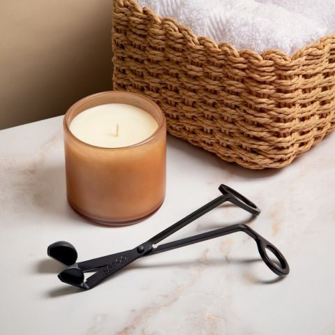 A luxurious LAFCO candle in a glass holder sits on a marble surface, accompanied by the sleek LAFCO Wick Trimmer and Snuffer. In the background, a woven basket holds neatly folded white towels, enhancing the elegant ambiance.