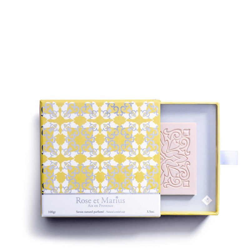 The ROSE ET MARIUS - SUNLIT ROSE WATER SCENTED SOAP GIFT BOX features a pastel pink square soap bar with an ornate, symmetrical floral design and carved swirls, displayed on a white background with shadows to the left and bottom.