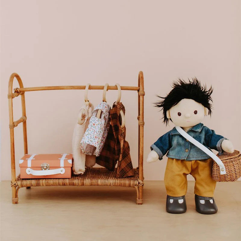 A Dinkum Doll from Olli Ella USA&#39;s Travel Togs collection, featuring dark hair and dressed in a denim jacket, mustard pants, and black shoes, stands on a wooden surface. Beside the doll is a teal See-Ya Suitcase with white trim and hardware. In the background, another larger teal suitcase is partially visible.