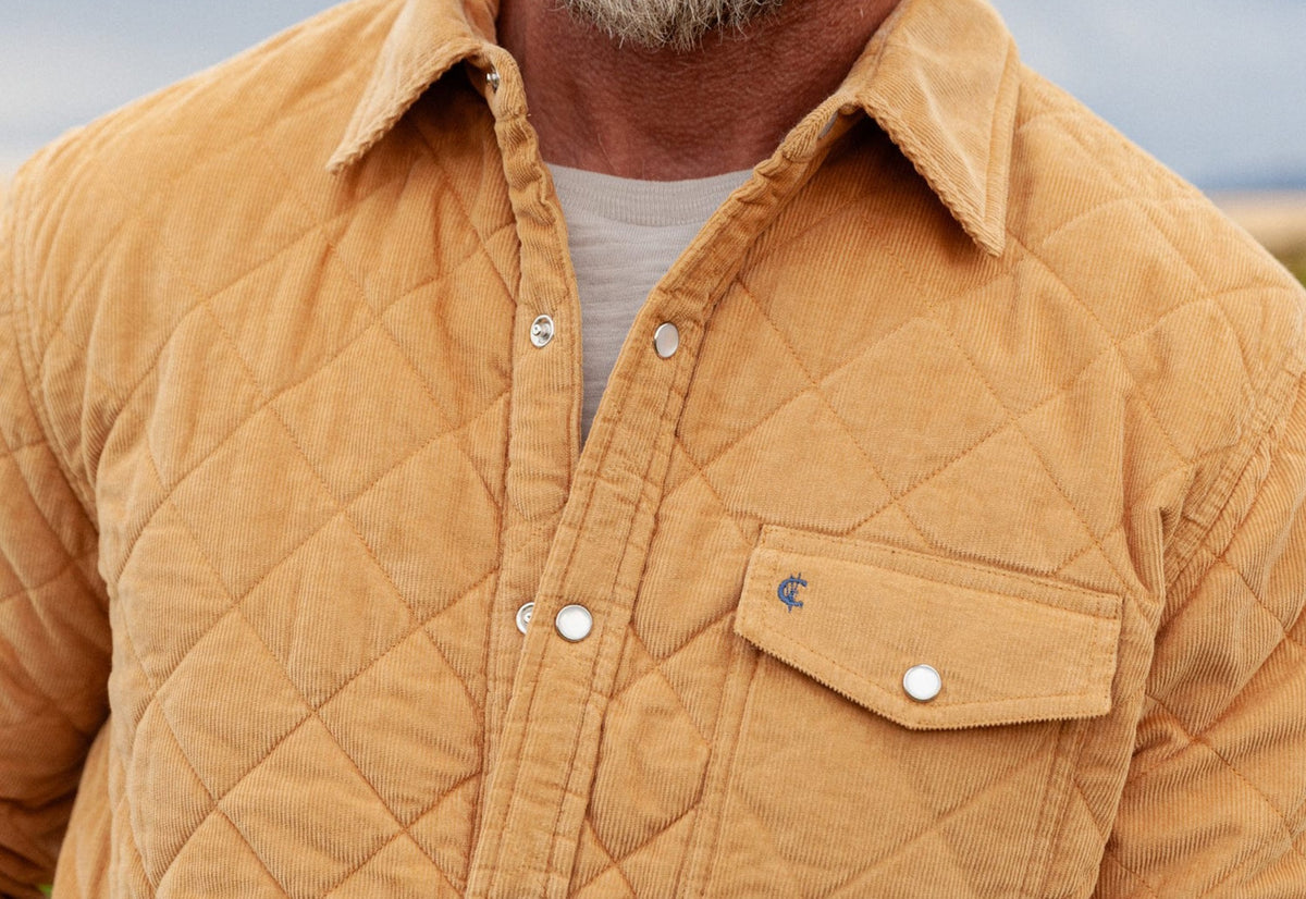 A man with short, curly hair and a beard wears the CRIQUET - CORDUROY QUILTED SHACKET by CRIQUET and blue jeans, standing against a white background while smiling and looking to his right.