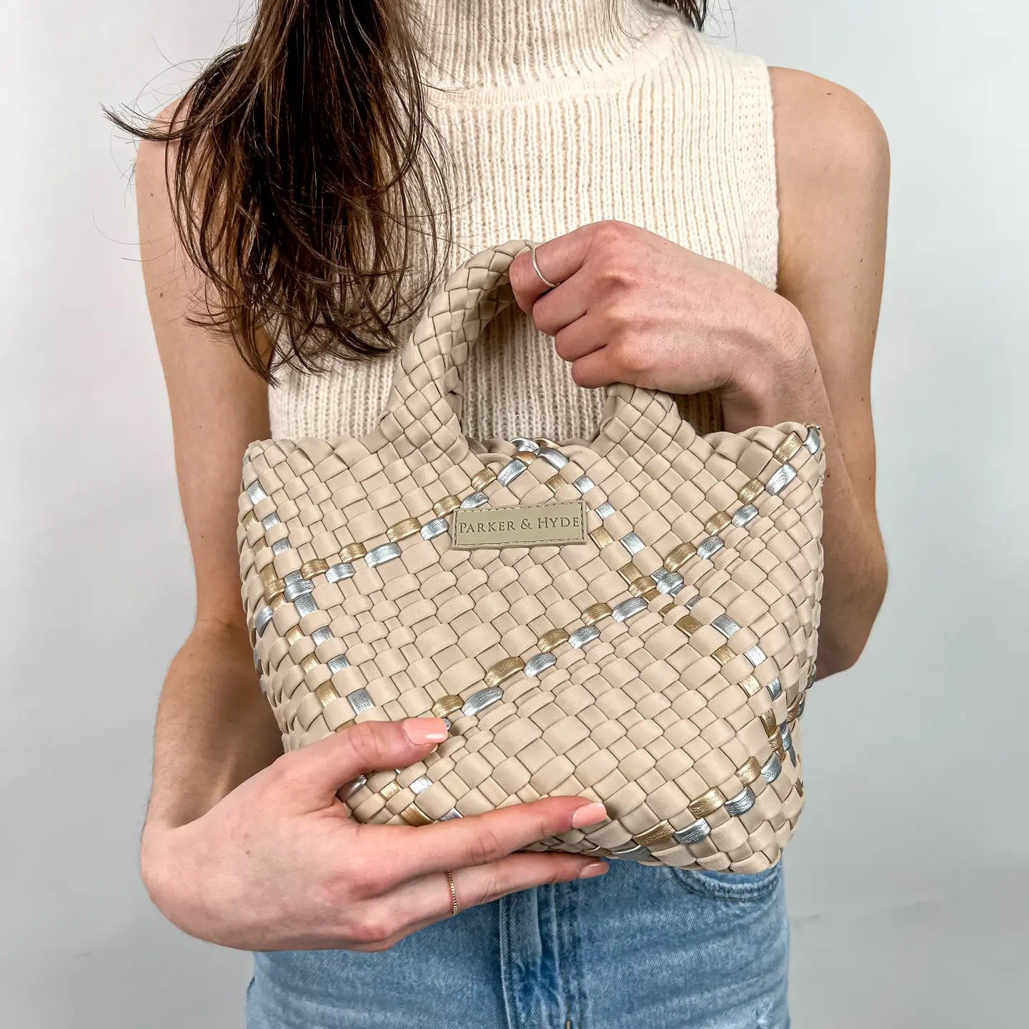 A woman with long brown hair, in a sleeveless cream knit top and blue jeans, holds a PARKER AND HYDE - MINI WOVEN TOTE BAG. The hand-woven tote by PARKER & HYDE features a subtle checkered pattern with light blue accents.