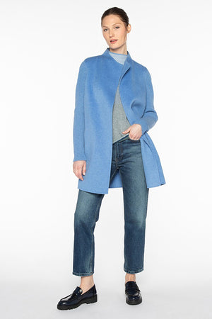 A woman stands against a plain white background, wearing the KINROSS CASHMERE - RIB SLEEVE COAT in blue over a grey top, paired with blue jeans and black loafers. She has her left hand in her pocket and her hair is tied back, looking relaxed and confident.