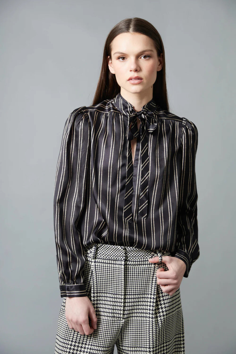 A woman with straight brown hair faces away in a HOPE FOR FLOWERS Tie Neck Blouse—black and white, vertically striped Tencel with puffed sleeves—paired with wide-leg plaid trousers. The plain gray background reflects sustainable style.