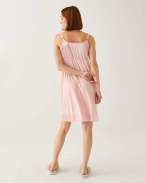 A woman with shoulder-length hair wears a light pink slip dress, like the elegant MOON DANCER PAJAMA DRESS by MER SEA, and fluffy slippers. She stands against a plain white background, looking directly at the camera with a neutral expression. Her arms are relaxed at her sides.