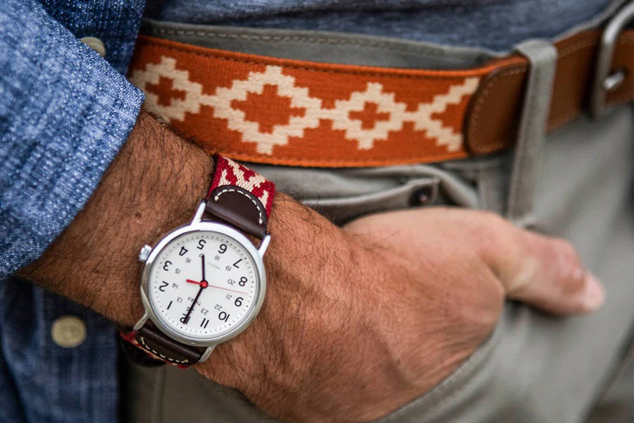 A man sporting a checkered shirt and the LA MATERA - TALAMPAYA WOVEN BELT stands by a classic teal car, hand on the door handle, ready to enter. The sunny scene unfolds in a residential area.