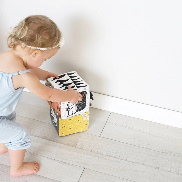 The WEE GALLERY - JUNGLE SOFT BLOCK by WEE GALLERY is a soft fabric cube perfect for stimulating a baby&#39;s developing senses. It features monochrome patterns and playful designs, including a cute monkey illustration on one side and a yellow leaf on another. Inspired by jungle life and animals, this toy boasts various textures and small loops on top for easy grasping.
