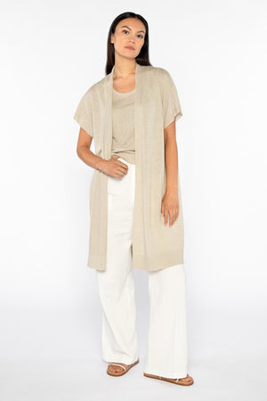 A woman with long dark hair, hands on hips, wears the KINROSS CASHMERE - LONG SHORT SLEEVE LINEN CARDIGAN in beige over white pants. Her back faces the camera against a plain white backdrop.