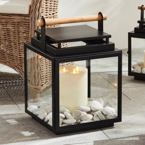 The ADWIN OUTDOOR LANTERN by NAPA HOME AND GARDEN is a black metal and glass outdoor lantern with a square design, featuring an exclusive teak handle on top. Inside the lantern, a lit white candle casts a soft, warm glow.