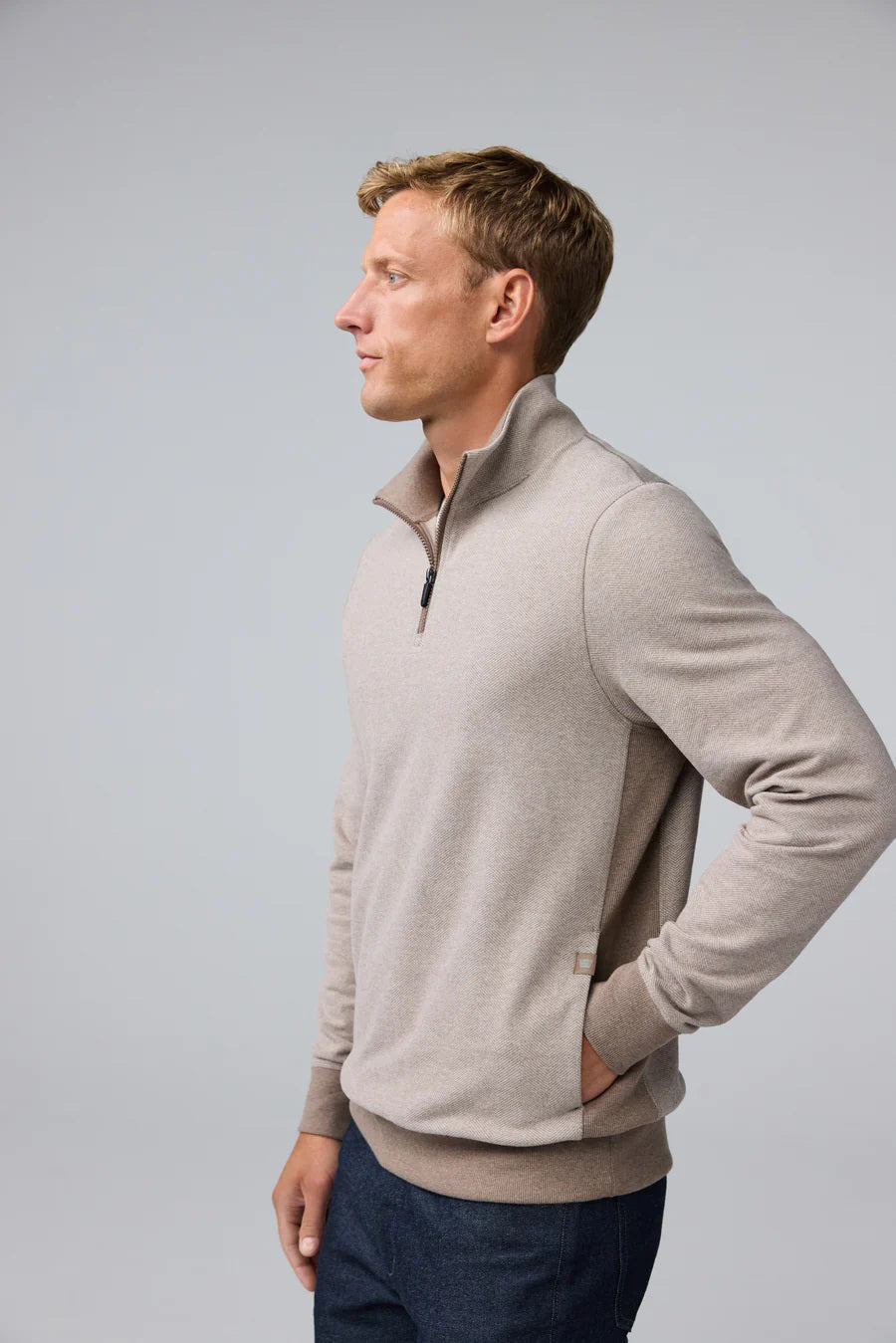 A man with short light brown hair stands in profile, wearing the MACK WELDON Ace 1/2 Zip Sweater in Herringbone and dark pants, one hand in his pocket, against a plain light gray background.