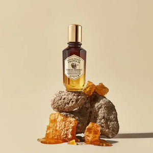 A bottle of KOSMIC KBEAUTY SKINCARE SKINFOOD ROYAL HONEY PROPOLIS ENRICH ESSENCE features a gradient design that transitions from dark brown at the top to golden yellow at the bottom, topped with a golden cap. The label emphasizes that it contains 63% black bee propolis and royal jelly extract for enhanced moisture and nutrition.