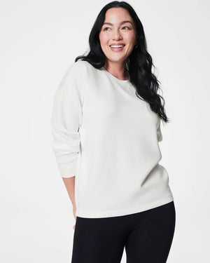 A person with long black hair stands facing slightly away from the camera, dressed in a SPANX AireEssentials Long Drew Sweater made of soft spacer fabric, complemented by black leggings and gray sneakers. The high-low hem on the sweater adds a touch of style against the plain white studio backdrop.