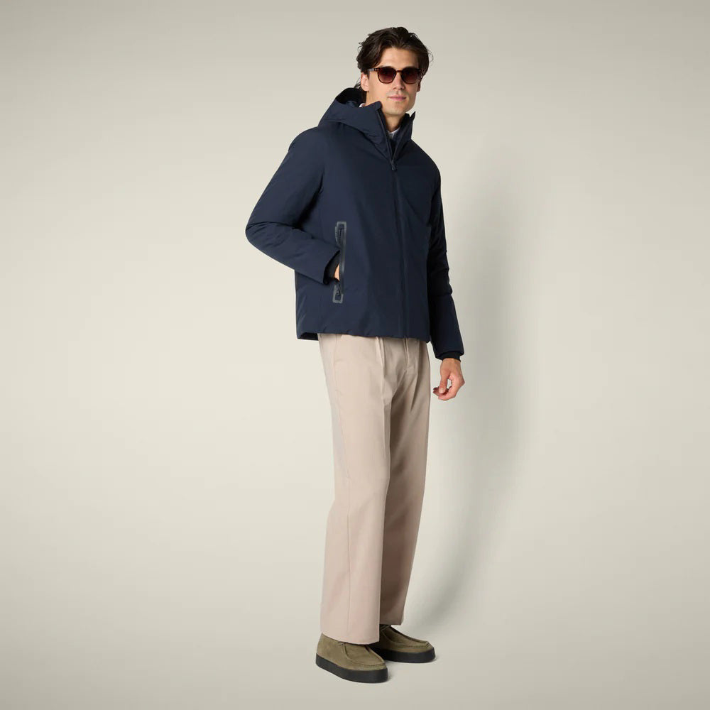 A man in sunglasses, beige wide-leg pants, and olive green shoes wears the SAVE THE DUCK - FLYNN HOODED JACKET by SAVE THE DUCK. He stands against a light background facing forward, with one hand by his side and one near his zipper.