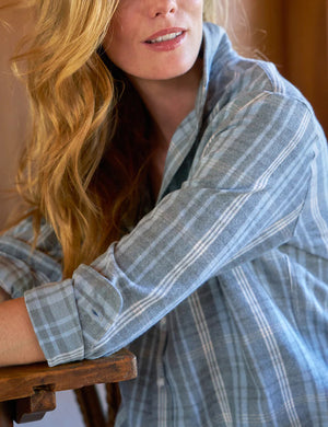 A person with long hair stands in a wooden room next to a window, wearing a relaxed fit FRANK & EILEEN EILEEN button-up shirt in grey cream blue plaid Italian flannel and white pants. Light floods the wicker chair and wooden walls.