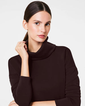 Dressed in the luxurious comfort of a SPANX AireEssentials Turtleneck Tunic, characterized by its black hue and lightweight fabric, a person stands with their back to the camera. Their elegantly tied up hair complements the matching black pants against a plain white backdrop.