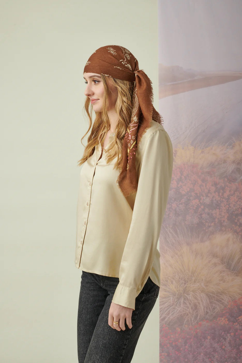 A young woman with light brown hair wears the STORIATIPIC Nelly Square Cashmere and Wool Embroidered Bandana Scarf, paired with a light gray top, standing before a softly colored nature-themed backdrop.