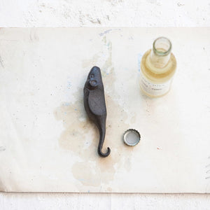 A compact, vintage-style cast iron bottle opener shaped like a mouse from Creative Coop rests on a white, slightly stained surface beside a half-full glass bottle labeled "Tonic Water" and its cap.