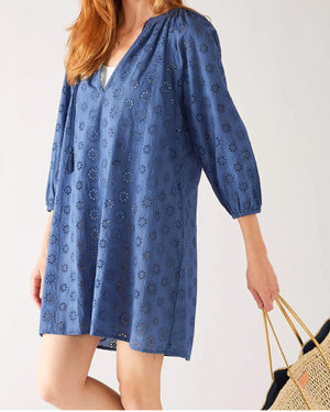 A person is seen wearing the MERSEA DAISY EYELET DRESS by MER SEA, a loose-fitting, knee-length, long-sleeved blue dress adorned with a daisy eyelet cutout pattern. The coastal elegance design features a V-neckline that reveals visible white fabric underneath. Made of 100% cotton, the person has one hand on their hip and the other hanging at their side.