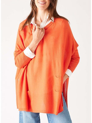 A woman with long hair wears a bright orange poncho over a white collared shirt and blue jeans. She is adjusting the poncho with her hand while smiling against a plain white background, showcasing a versatile look that can easily replace your favorite MERSEA Catalina Crewneck Sweater for any occasion.