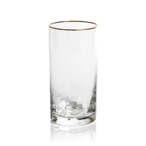 Two textured glass tumblers with gold rims sit on a sparkling surface. The taller, cylindrical tumbler, identified as the NEGRONI HAMMERED HIGHBALL GLASS by ZODAX, has a capacity of 560 ml, while the shorter and wider one holds less. Blurred green bottles and a white bowl in the background enhance the subtle and elegant atmosphere.