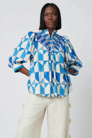 A woman with shoulder-length black hair wears the Hope for Flowers - Smocked Blouse, which features a geometric pattern in shades of blue and white. This sustainable fabric blouse by Hope for Flowers boasts a high collar with buttons and puffed sleeves. She pairs it with light-colored pants that have decorative elements. The backdrop is a plain light gray.