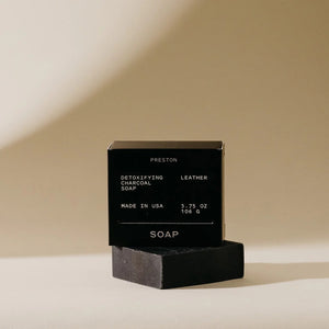 A minimalist arrangement of geometric shapes, featuring a white sphere perched on two black rectangular blocks stacked vertically, with a white cube placed at the base. The objects cast soft shadows against a textured wall, reminiscent of the simplicity seen in PRESTON's premium bar soap packaging designs.