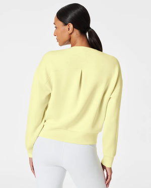 A woman stands confidently against a plain white background, wearing the SPANX Aireessentials Crew Neck Pullover and matching sweatpants made from luxurious comfort spacer fabric. She has her hair pulled back and her left hand is raised slightly. She smiles gently with her lips closed.