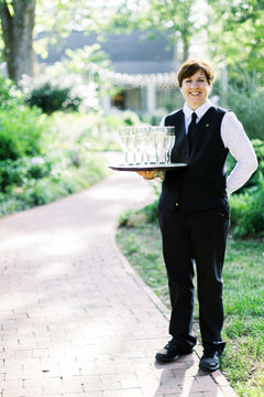 A smiling server in a black vest and white shirt stands on a brick path in a garden, holding a tray with several empty champagne flutes. Lush greenery and a building appear in the background.