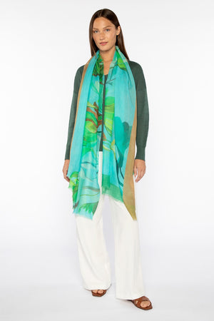 A person stands against a white background, wearing a green top, white pants, and brown sandals. They drape the KINROSS Acacia Print Silk and Cashmere Scarf—a luxury accessory with blue and green leaf designs—around their neck.