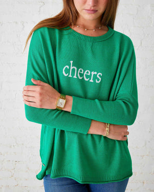 A woman in a versatile green MERSEA - CHEERS CATALINA CREWNECK SWEATER, featuring the word "cheers," stands against a white brick wall, smiling with her hands at her sides. This staple piece from MER SEA pairs perfectly with blue jeans and a gold bracelet.