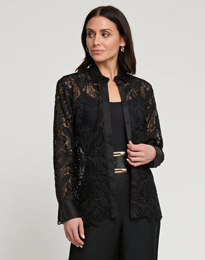 A woman with dark hair stands against a plain background, wearing the HINSON WU - MARGOT LACE SHIRT JACKET, featuring intricate lace detailing and satin trim. She has elegantly paired it with dark pants, her profile turned slightly sideways as she offers a subtle smile.