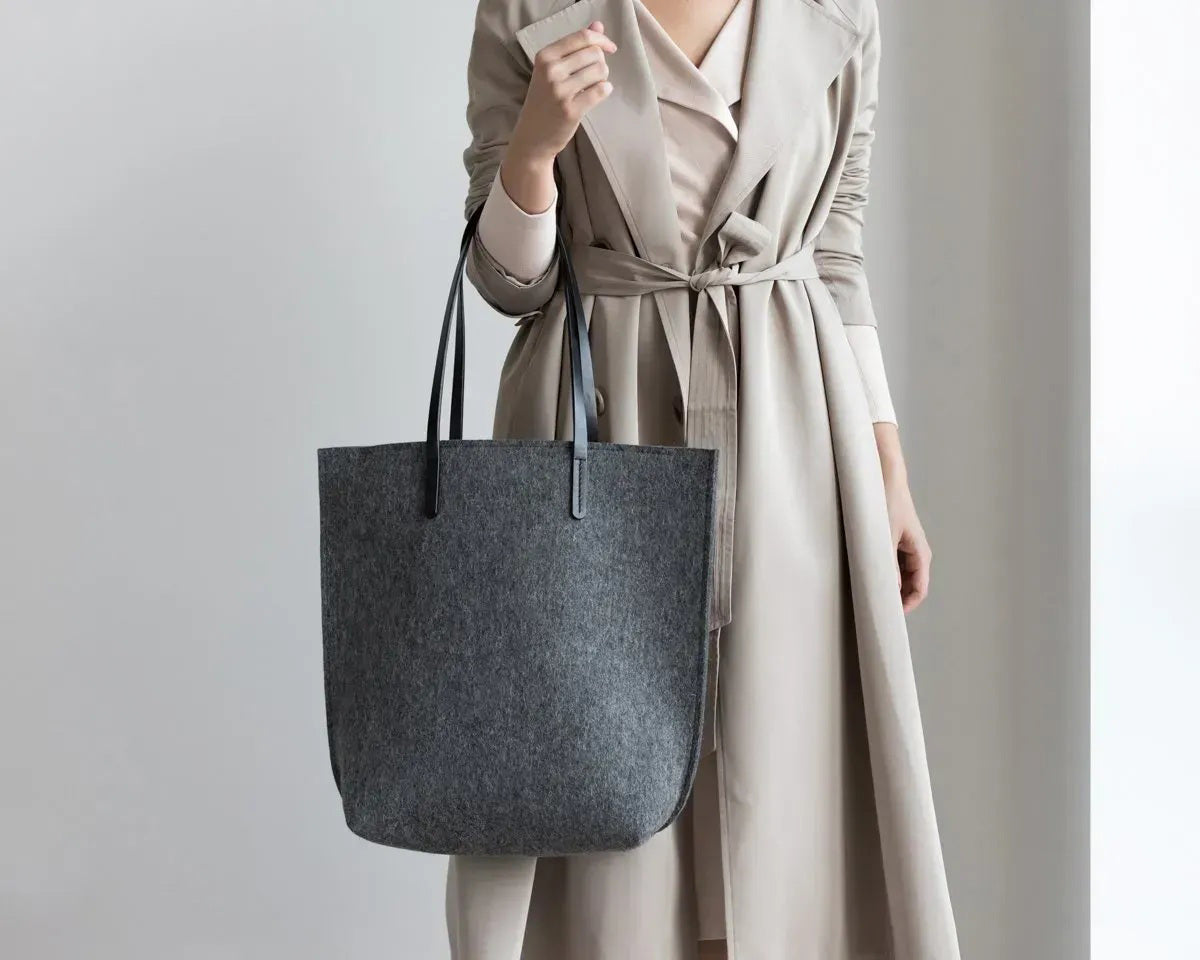 Close-up of hands in a beige knit sweater holding the WOOLBERRY WOOL FELT LONG TOTE BAG, featuring black leather straps and silver hardware.
