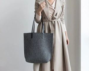 Close-up of hands in a beige knit sweater holding the WOOLBERRY WOOL FELT LONG TOTE BAG, featuring black leather straps and silver hardware.