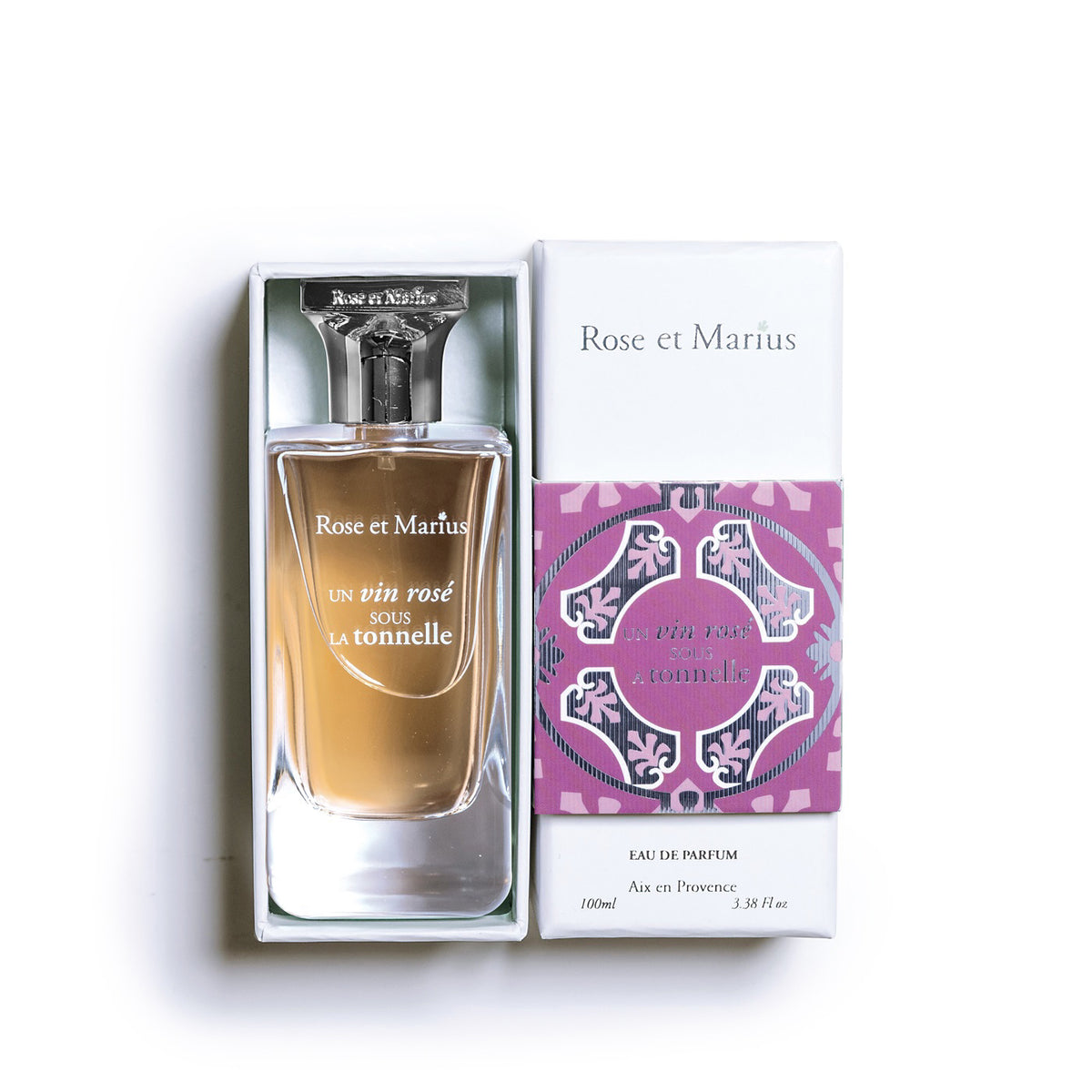A clear glass bottle of ROSE ET MARIUS - A ROSE WINE UNDER THE ARBOR Eau de Parfum 100ml, known for its fruity woody scent, with a patterned square cap beside it on a white background. The bottle casts a soft shadow.