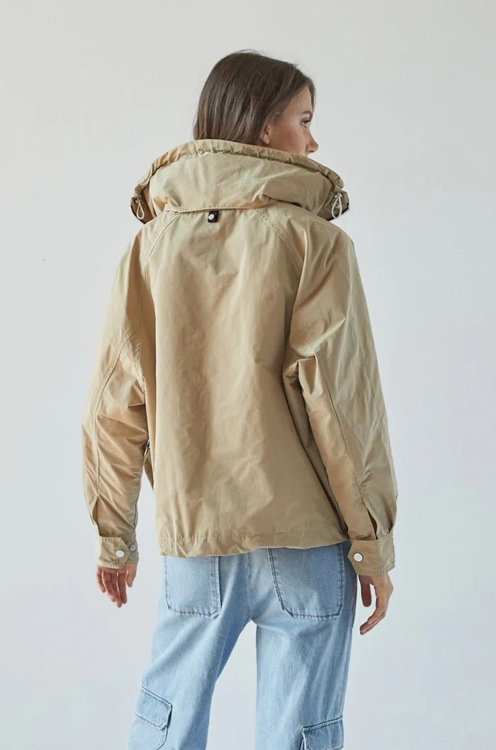 A person with long brown hair, wearing the ADROIT ATELIER - RYDER BOMBER JACKET in beige, featuring a high collar and hidden hood, along with blue jeans is standing with their back to the camera. The background is plain and light-colored.