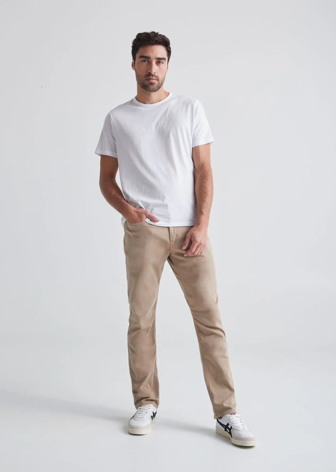 A man with short dark hair and a trimmed beard stands against a white background, wearing a white t-shirt, white sneakers with black details, and DUER&#39;s DU/ER NO SWEAT RELAXED TAPER PANT in an athletic fit. One hand is in his pocket.