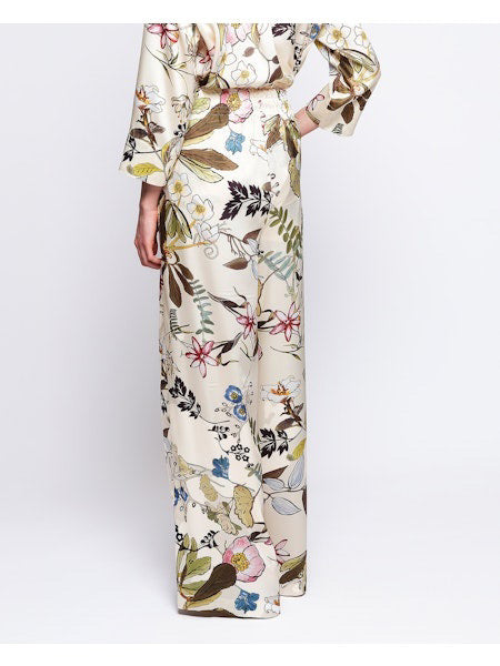 The woman showcases the MIRTO 1956 Botanical Floral Print Fluid Pants with an elastic waistband, complemented by a matching wide-brimmed hat and floral outfit, against a plain background.