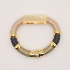 The HOLST & LEE - Luxe Colorblock Bracelet showcases sections of black, teal, and red thread, accentuated by gold-tone rings and a central rhinestone-studded bead. The gold-plated clasp is engraved with "HOLST" and "LEE" labels.