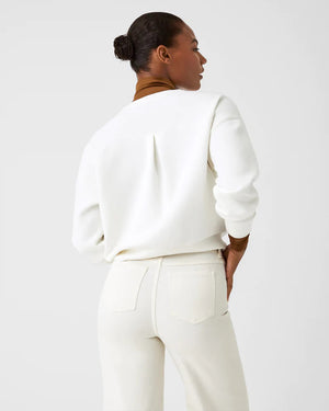 A woman stands confidently against a plain white background, wearing the SPANX Aireessentials Crew Neck Pullover and matching sweatpants made from luxurious comfort spacer fabric. She has her hair pulled back and her left hand is raised slightly. She smiles gently with her lips closed.