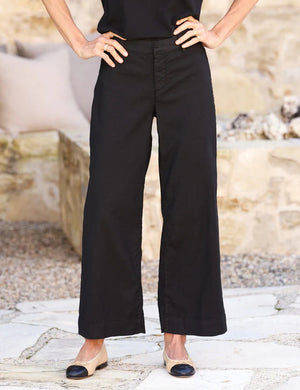 A person is standing with hands on hips, wearing the FRANK & EILEEN - WEXFORD WIDE LEG LINEN PANT IN BLACK and beige flats with black toe caps. They are outdoors, with a blurred stone wall and ground in the background.