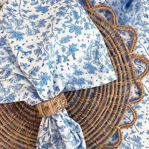 The CHRISTINA DICKSON HOME - WHIMSY FLORAL COTTON NAPKIN, an artisan-crafted blue and white floral fabric made in India, is tied in a knot over a circular rattan mat. Its delicate leaf and flower design complements the mat's textured pattern.