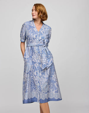 A person with shoulder-length hair wears the HINSON WU Luca Elbow Sleeve Lace Print Dress, a blue and white breathable dress featuring short sleeves, a belted waist, and a hand in a pocket against a plain background.