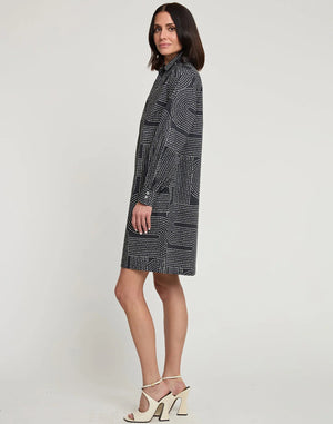 A woman with shoulder-length dark hair is wearing the new HINSON WU Vicky Long Sleeve Dress with Saddle Stitch Print, a knee-length outfit featuring a geometric dot pattern in shades of grey and white. Paired with white high-heeled sandals, she stands against a plain, off-white background.