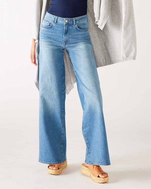 A person models the MERSEA - KATE WIDE LEG DENIM by MER SEA, light blue high-waisted jeans made from recycled cotton, paired with a dark top and gray cardigan against a neutral backdrop. Tan wedge sandals complete the look, emphasizing the stylish denim from the waist down.