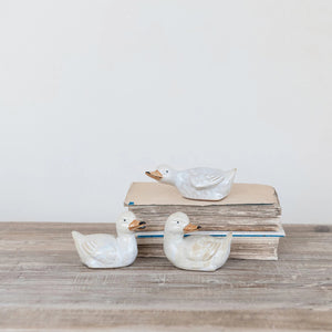 Three STONEWARE DUCKLING figurines by CREATIVE COOP sit on and in front of vintage books with worn edges, set against a plain white backdrop on a wooden surface, evoking nostalgia in their ceramic vintage reproduction form.