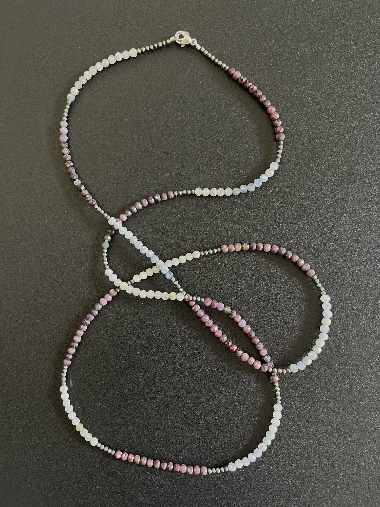 The ANN LIGHTFOOT - LONG STRAND SAPPHIRE, AQUAMARINE AND PYRITE NECKLACE showcases small, round beads in shades of pink, white, and purple elegantly intertwined on a dark surface. Accentuated by hints of aquamarine, this piece is completed with a clasp at one end.