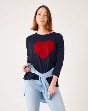 Confident in a MER SEA Catalina long-sleeve navy slub tee with a red heart, they style it effortlessly with a light blue denim shirt tied around their waist. The oversized fit pairs perfectly against the plain white background, highlighted by their light brown hair.